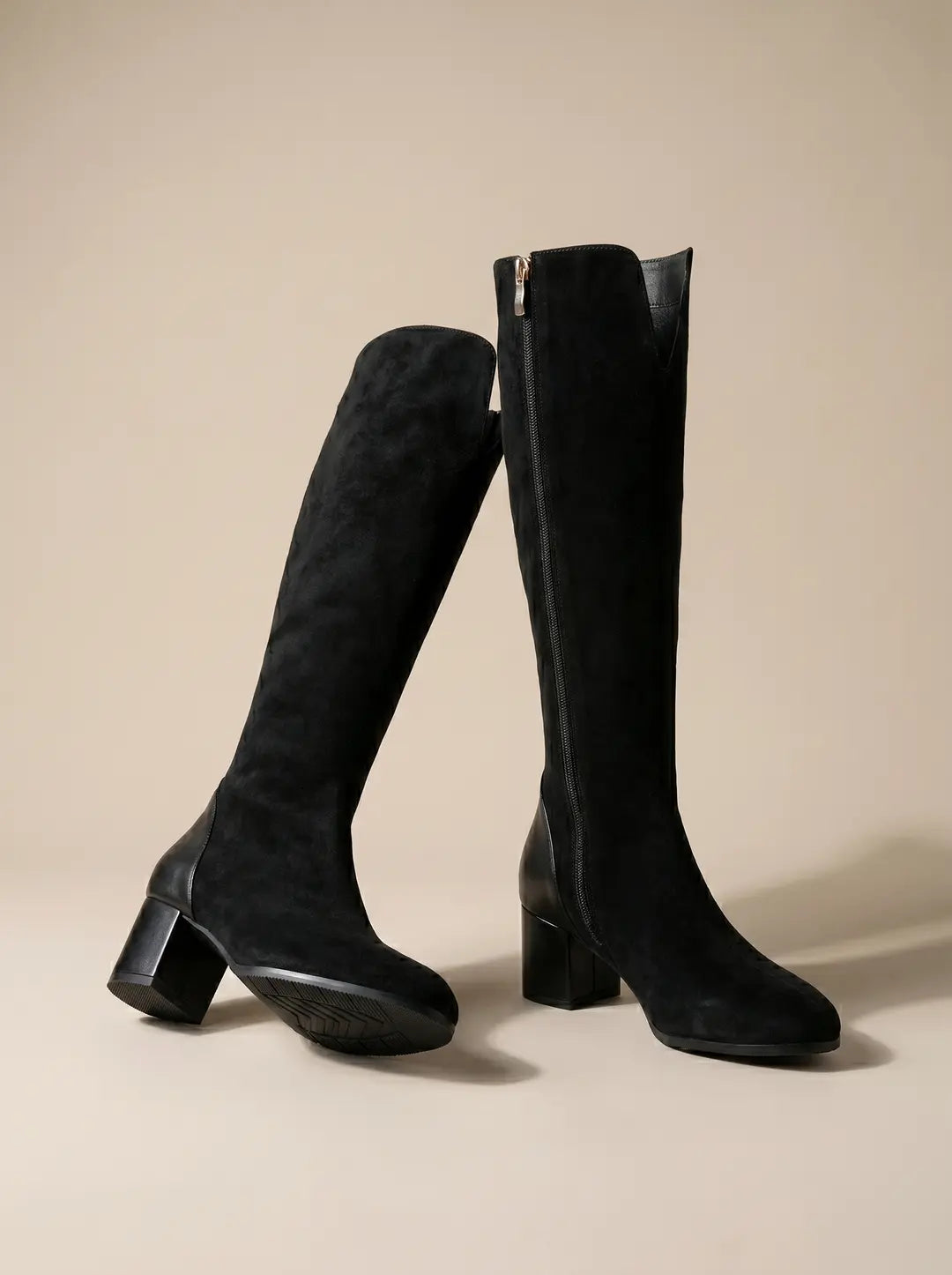 Par de botas altas negras de ante con detalles de cuero, tacón de bloque y cremallera lateral, sobre un fondo liso beige claro.