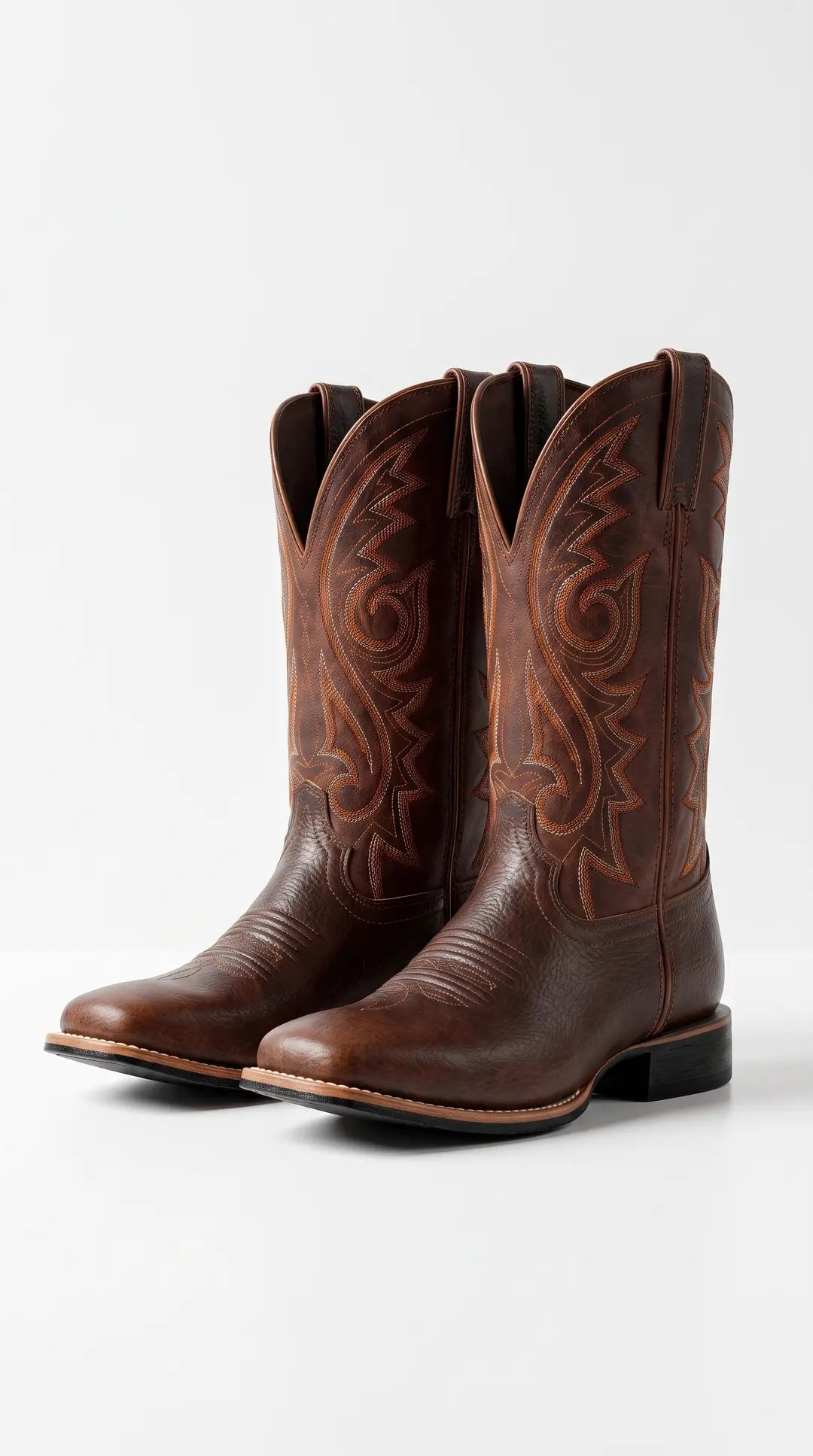 Par de botas vaqueras de cuero marrón con costuras decorativas en las cañas, puntera cuadrada y tacón apilado, sobre un fondo blanco.