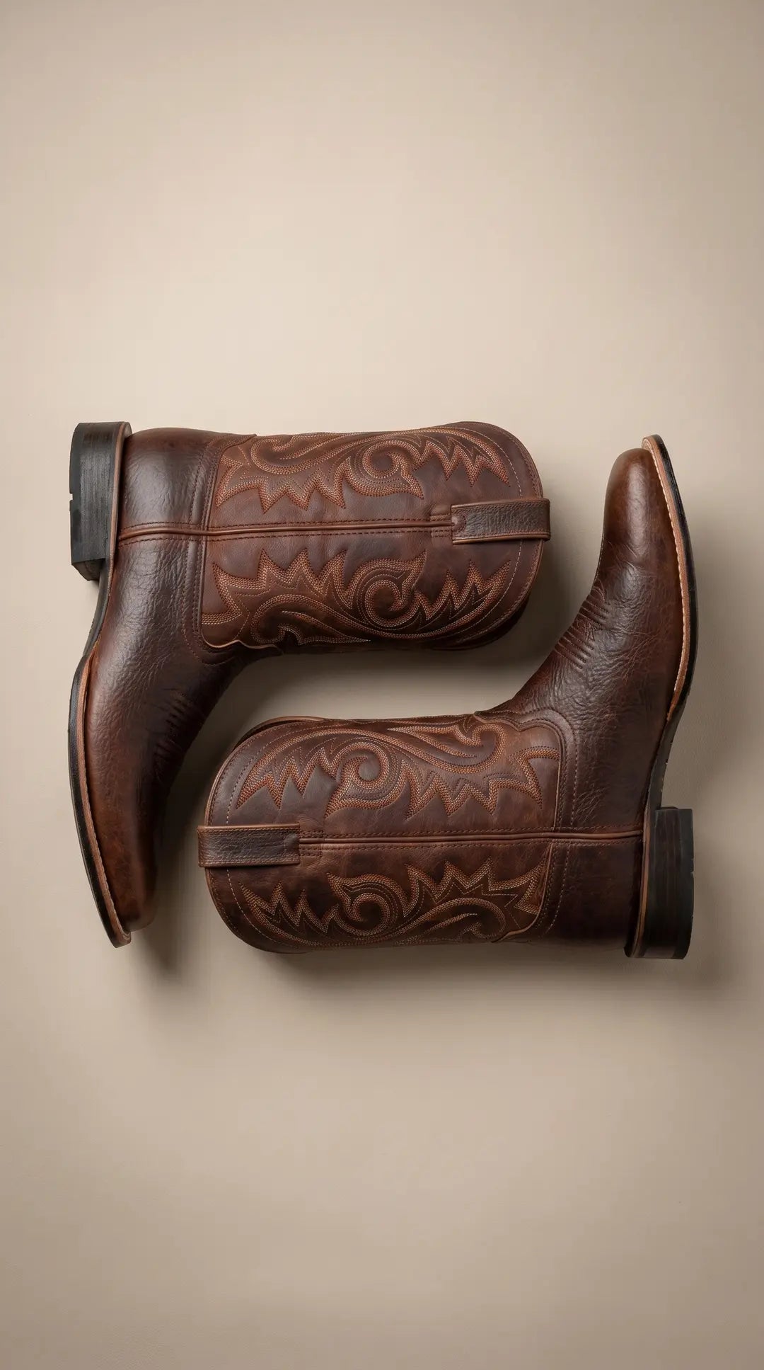 Dos botas vaqueras de cuero marrón oscuro con intrincados bordados, dispuestas horizontalmente sobre una superficie beige clara.