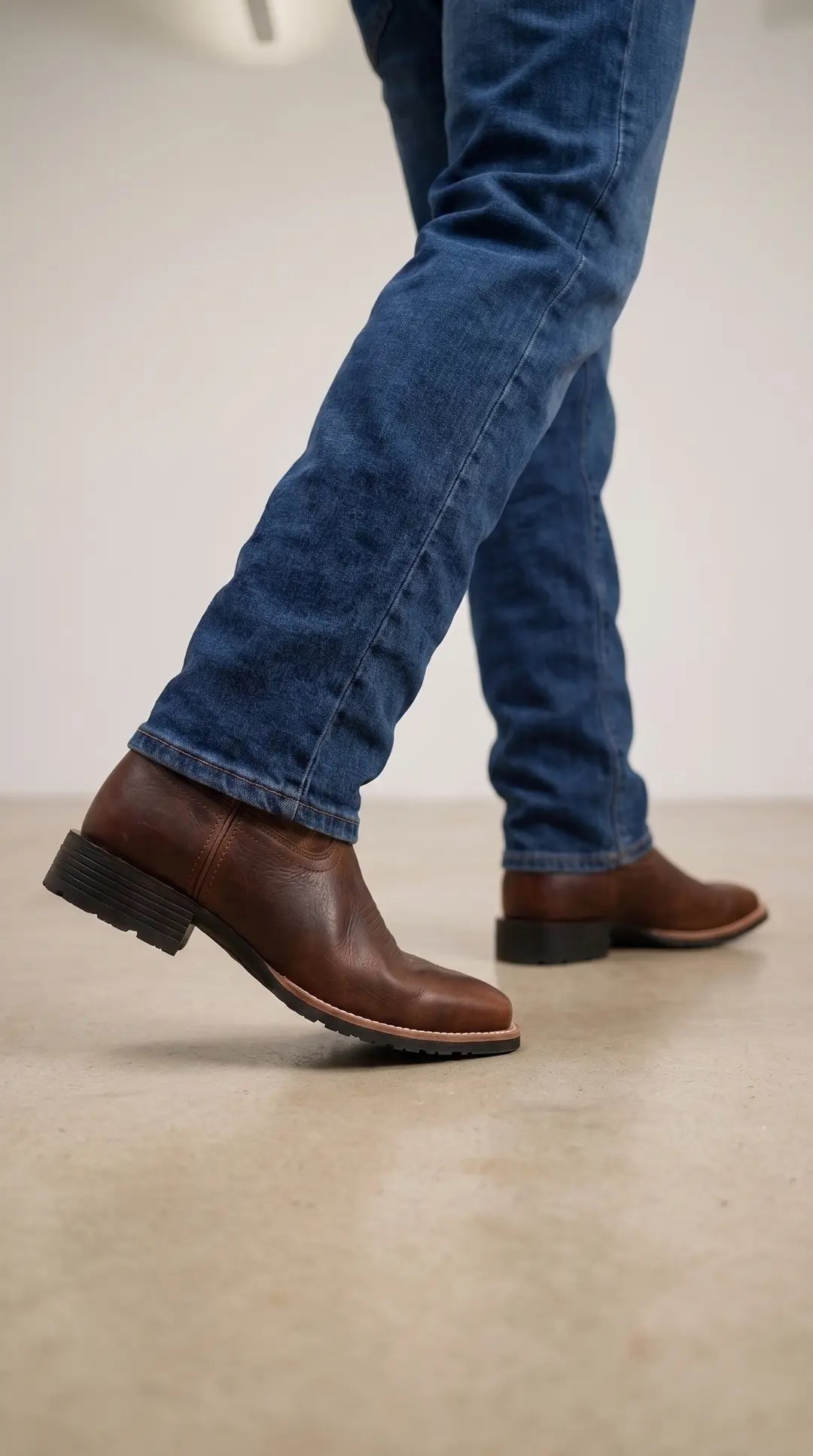 Piernas de una persona con jeans azules y botas de cuero marrón oscuro, con tacones negros y costuras claras visibles en la suela, caminando sobre un suelo claro.