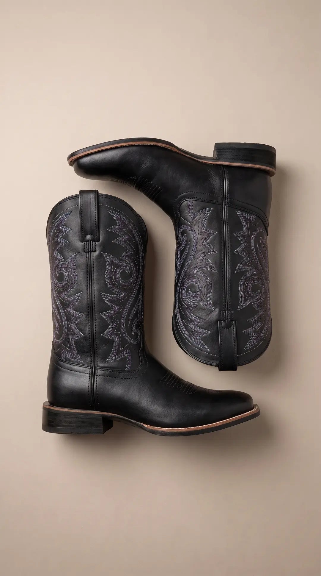 Dos botas vaqueras negras con bordados morados y blancos en la caña, dispuestas planas sobre un fondo beige claro.