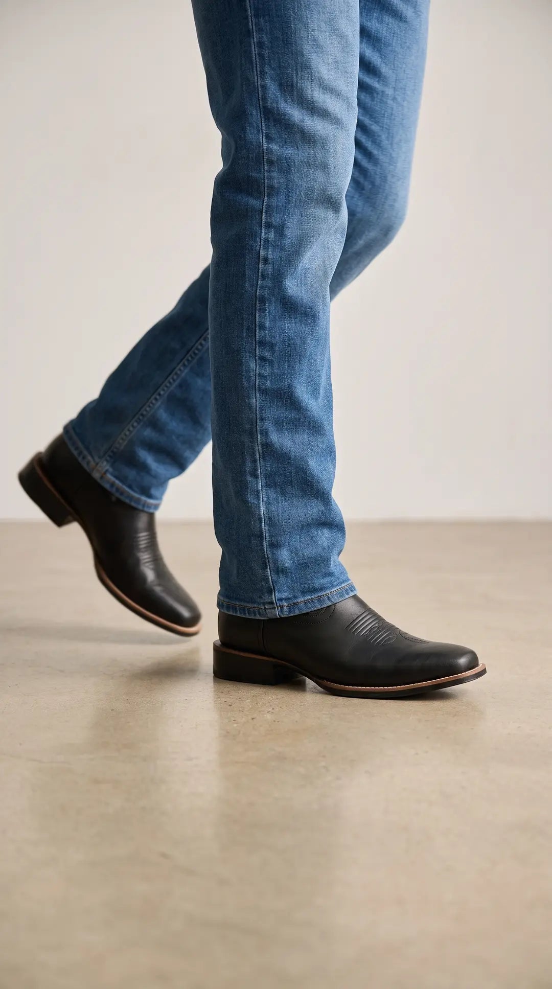 Piernas de una persona con jeans azules y botas vaqueras negras. Las botas tienen punta cuadrada, tacón bajo y suela de borde marrón claro, caminando sobre un piso claro.