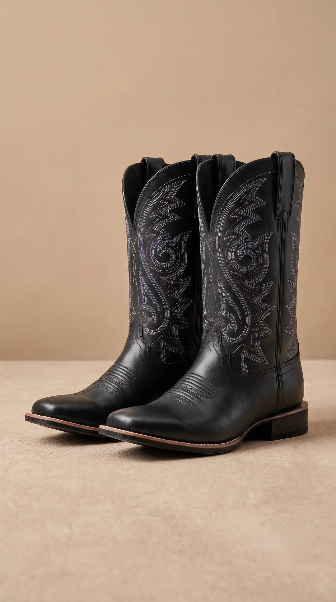 Par de botas de vaquero negras con puntera cuadrada y bordados decorativos en tonos morados y blancos en la caña, sobre una superficie beige.