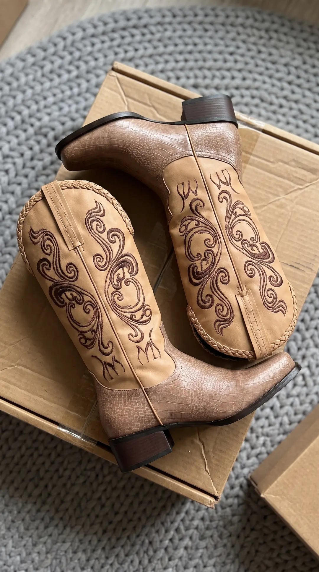 Un par de botas vaqueras de color beige con textura de piel de cocodrilo en la parte inferior y tacones marrones. La caña de la bota es de cuero liso con bordados de espirales en marrón y un borde superior trenzado. Las botas están sobre una caja de cartón encima de una alfombra de punto gris.