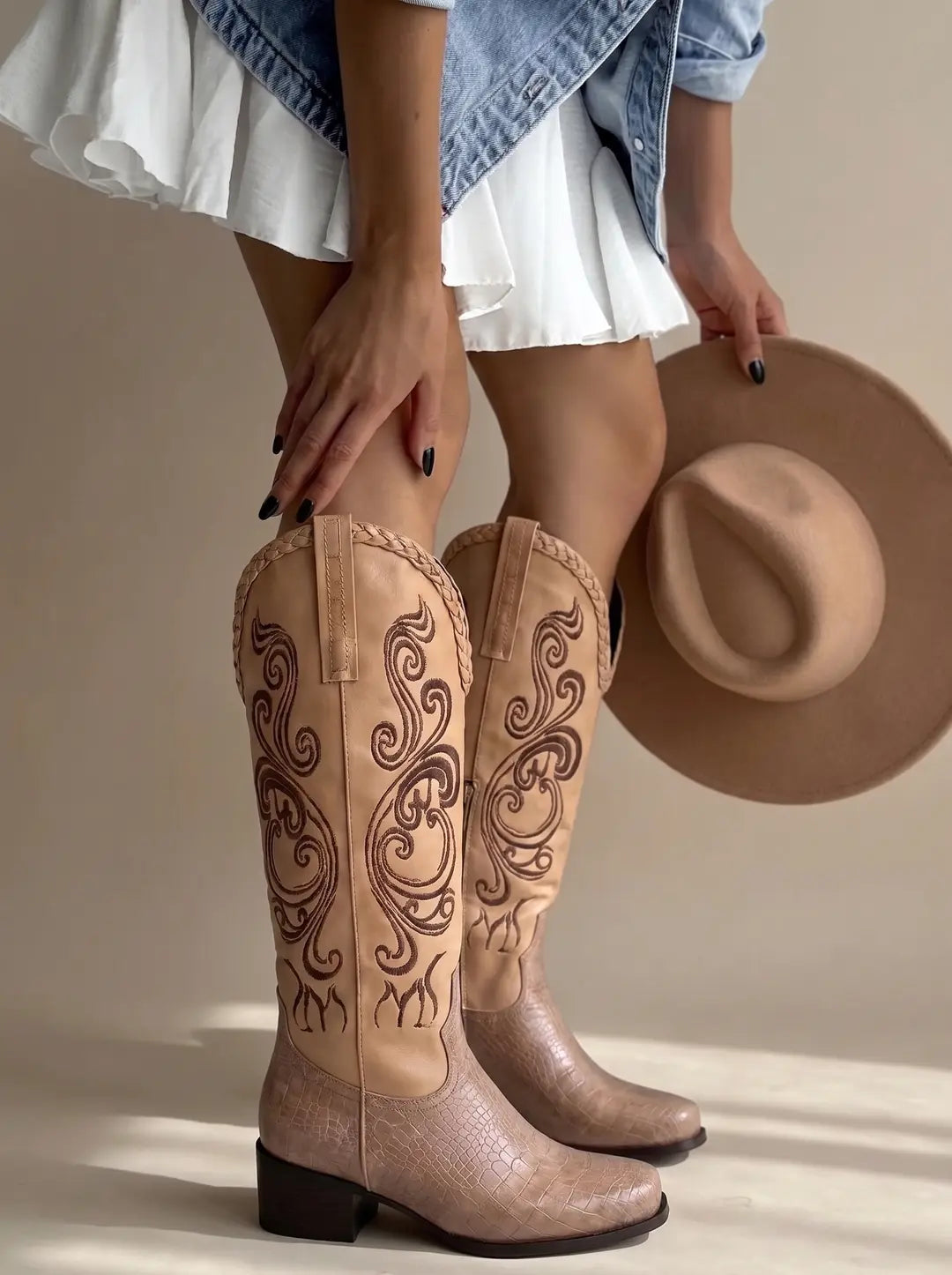 Botas vaqueras altas de color beige con bordados marrones en la caña y textura de cocodrilo en el empeine. Una persona sostiene un sombrero de fieltro beige a la derecha.