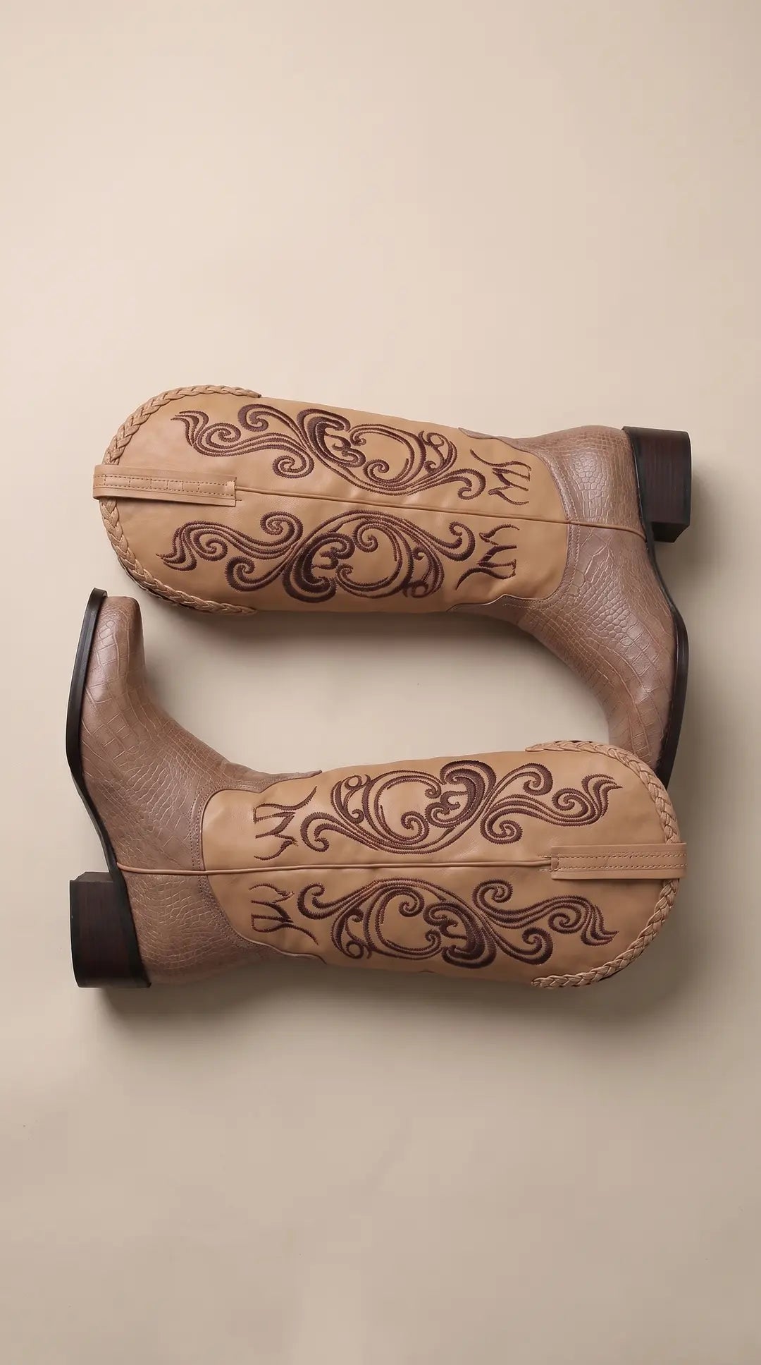 Par de botas de vaquero con caña beige y bordados marrones, con parte inferior y tacón en textura de piel de cocodrilo color topo, sobre un fondo liso.
