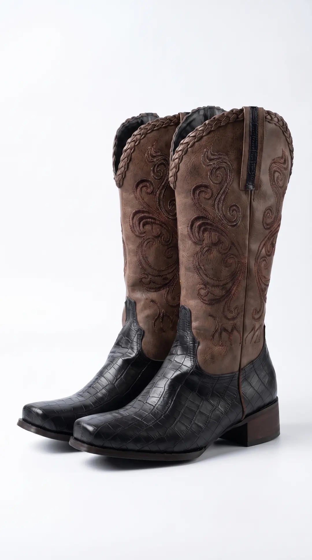 Par de botas vaqueras de dos tonos. La parte inferior es negra con textura de piel de cocodrilo y la caña superior es marrón con bordados en espiral y un borde trenzado.