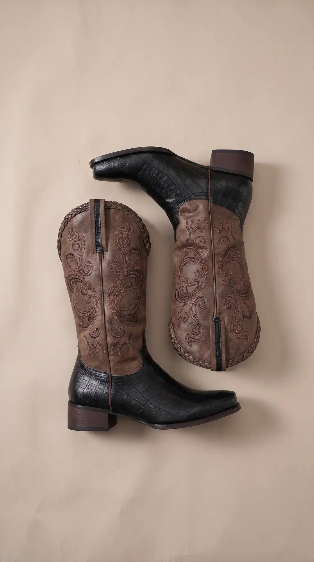 Par de botas vaqueras bicolores sobre un fondo claro. La parte superior es marrón con bordados y un borde trenzado, y la parte inferior es negra con patrón de cocodrilo. Tienen tacones oscuros.