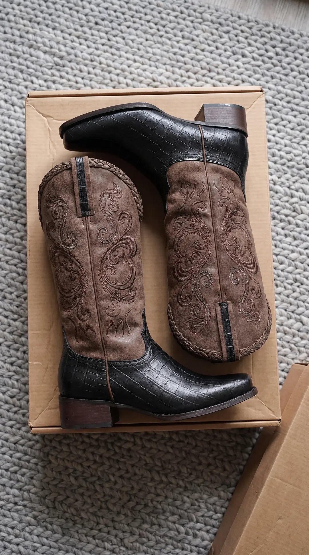 Par de botas vaqueras bicolor con la parte del pie negra texturizada y la caña marrón con bordados ornamentales. Las botas tienen un borde superior trenzado y están sobre una caja de cartón encima de una alfombra gris.