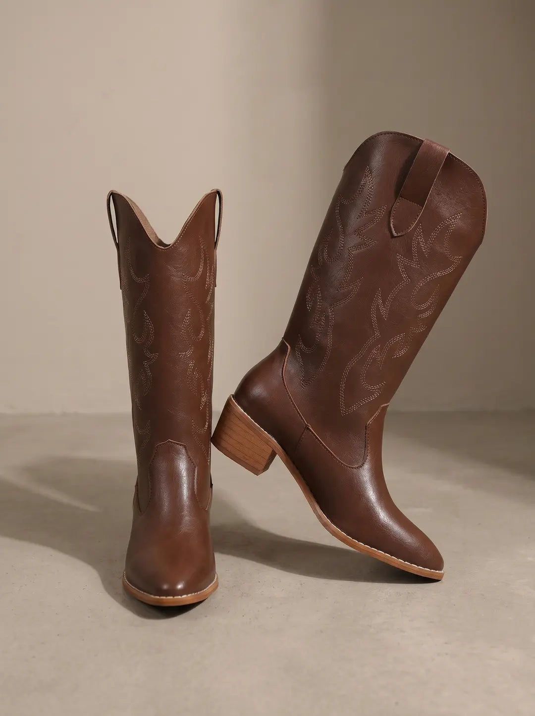 Dos botas vaqueras de color marrón oscuro con puntera puntiaguda, tacón de bloque y costuras decorativas en la caña. Una bota está de pie y la otra inclinada.
