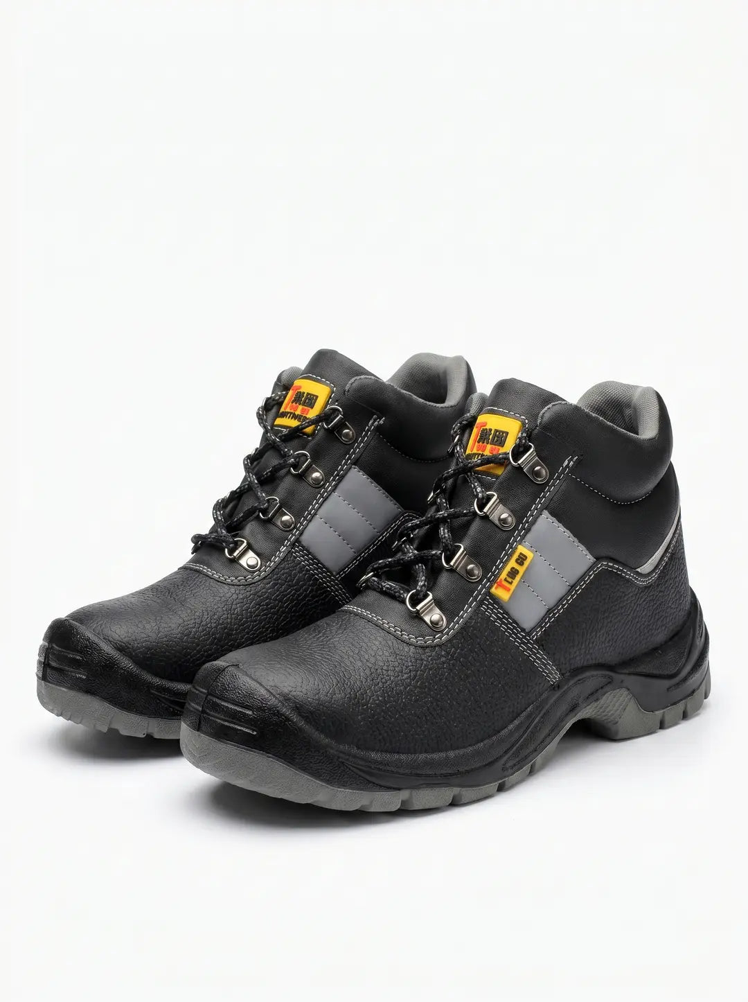 Un par de botas de seguridad negras con suelas grises, cordones negros, ojales metálicos y detalles de costura blanca y franjas grises laterales. Cada bota tiene una etiqueta amarilla y roja cerca de los cordones.