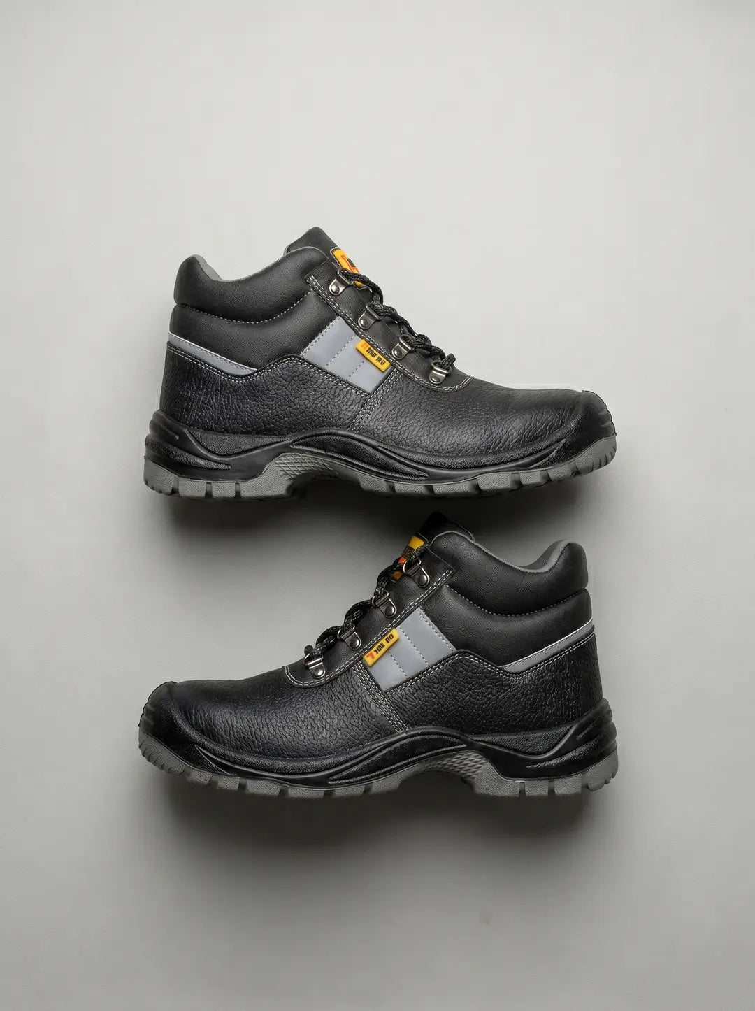 Dos botas de trabajo negras con suelas y detalles laterales grises, cordones negros y ojales metálicos. Las botas tienen una textura granulada y están dispuestas una encima de la otra, orientadas a la izquierda, sobre un fondo gris claro.
