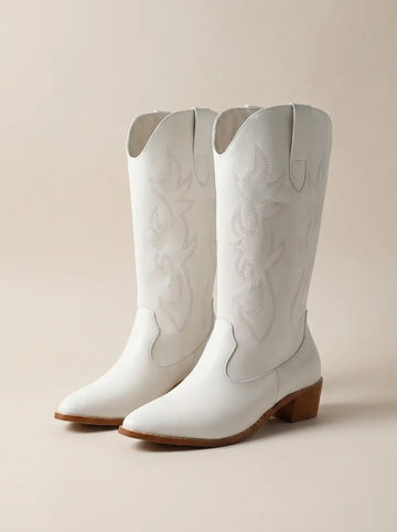 Par de botas vaqueras blancas con bordados decorativos, tacones y suelas marrones, sobre un fondo beige claro.