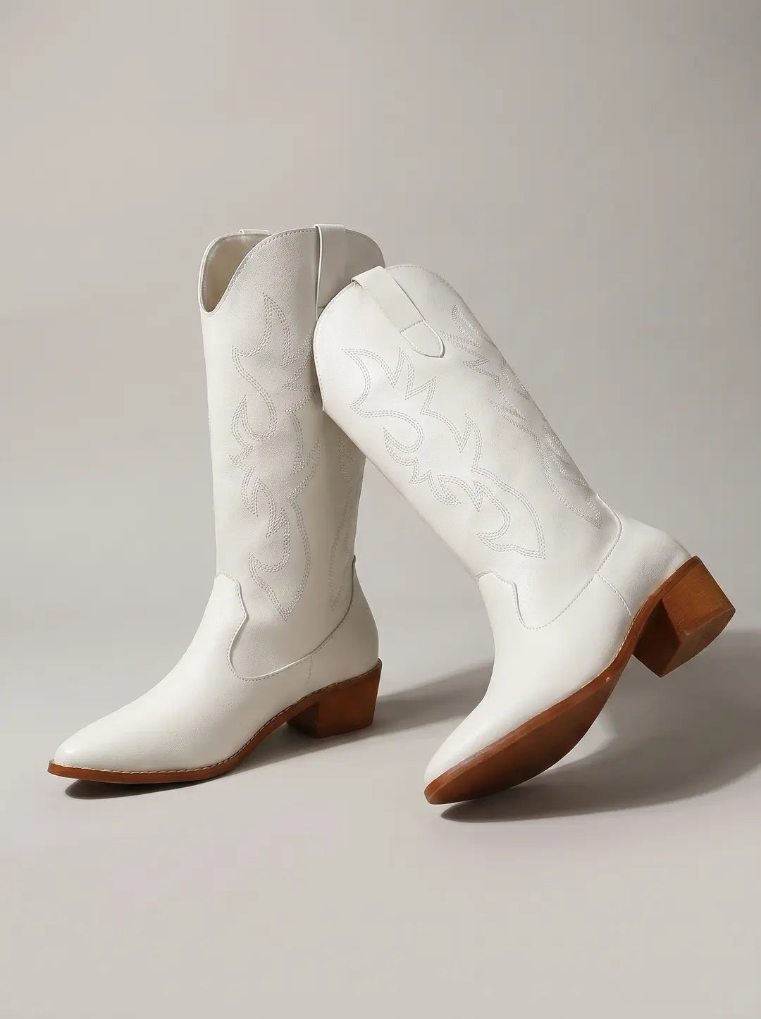 Dos botas vaqueras blancas. Una está de pie y la otra ligeramente inclinada. Ambas botas tienen detalles de costuras decorativas en la caña y el empeine, puntas puntiagudas y tacones bajos y suelas de color marrón.