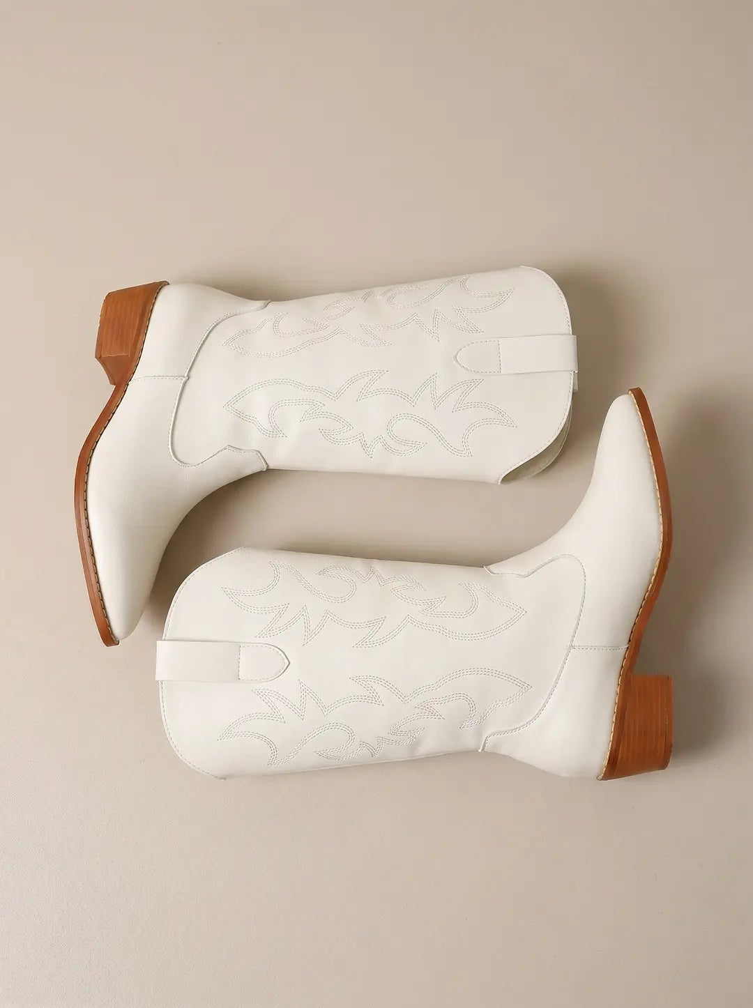 Dos botas de vaquero blancas con costuras decorativas y tacones bajos de color madera, dispuestas en diagonal sobre un fondo beige liso.