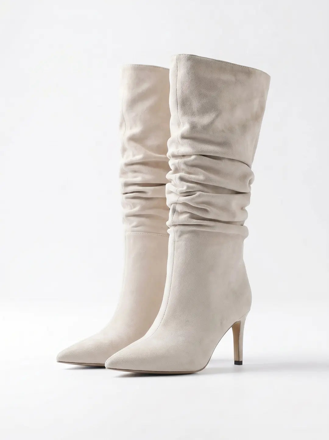 Un par de botas de tacón alto de ante en color beige claro o crema, con caña arrugada y punta afilada, sobre un fondo blanco.