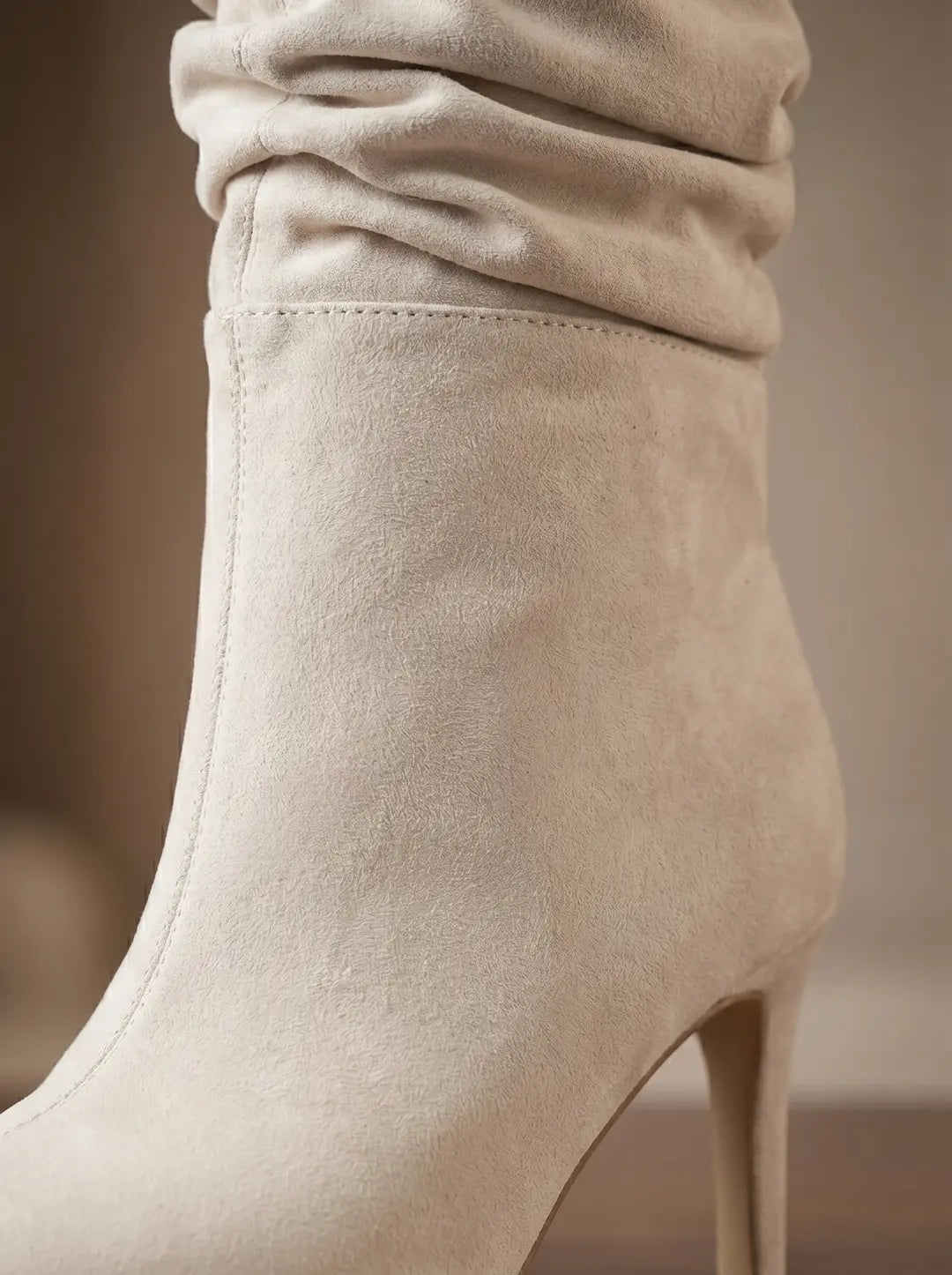 Bota color crema con textura de gamuza, caña fruncida y tacón de aguja. Se aprecian detalles de costura en la parte superior del calzado.