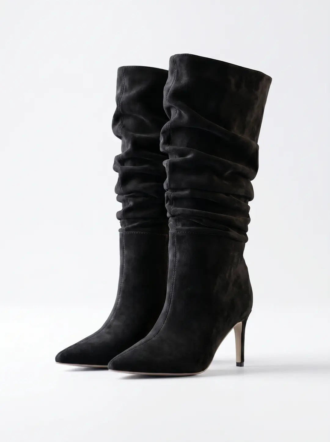 Dos botas de mujer negras de ante, con caña alta arrugada, puntera afilada y tacón de aguja en tono claro, sobre un fondo blanco.