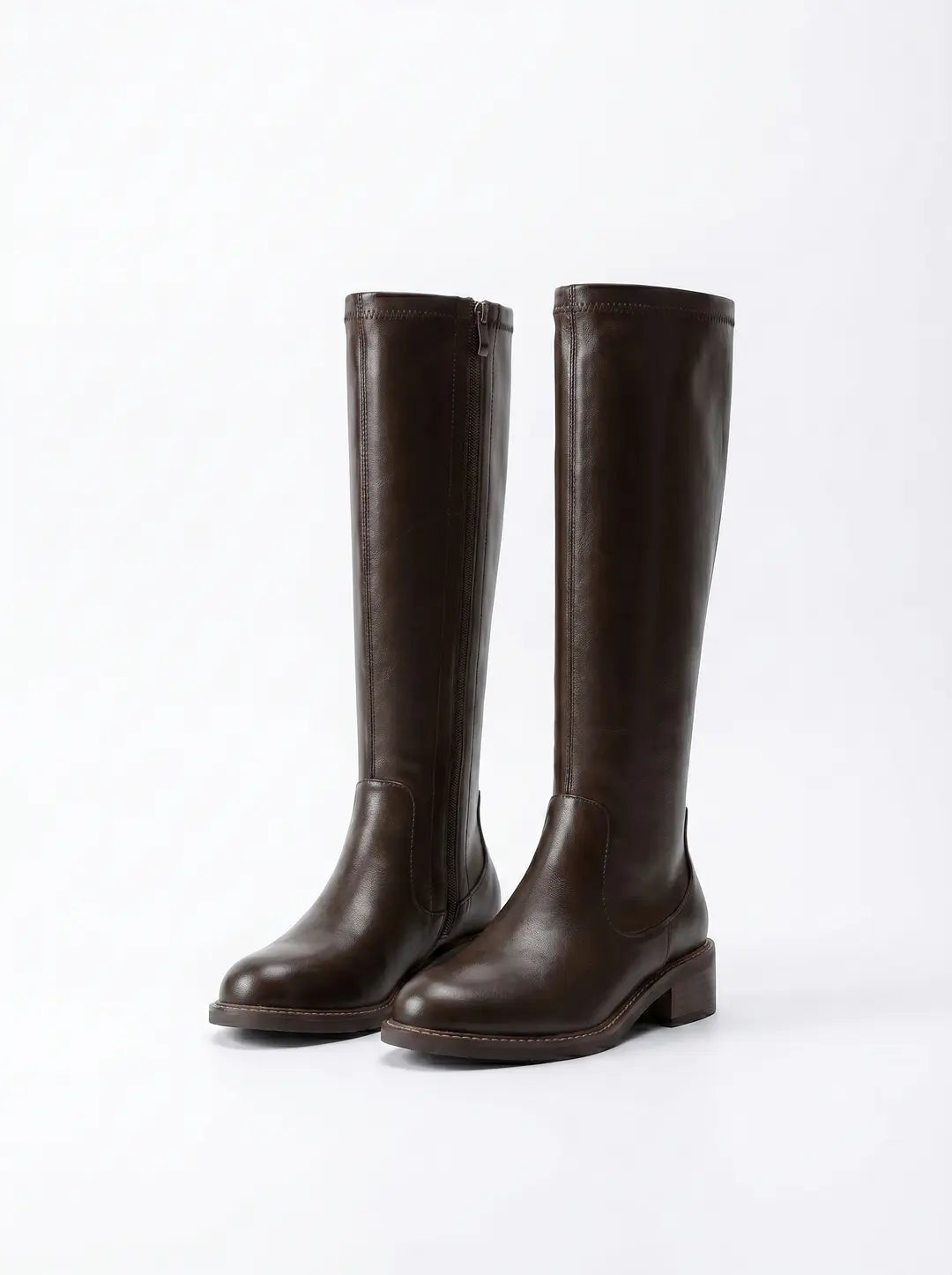 Dos botas altas hasta la rodilla de color marrón oscuro con textura lisa y tacón bajo, una de ellas con cremallera lateral, sobre un fondo blanco.