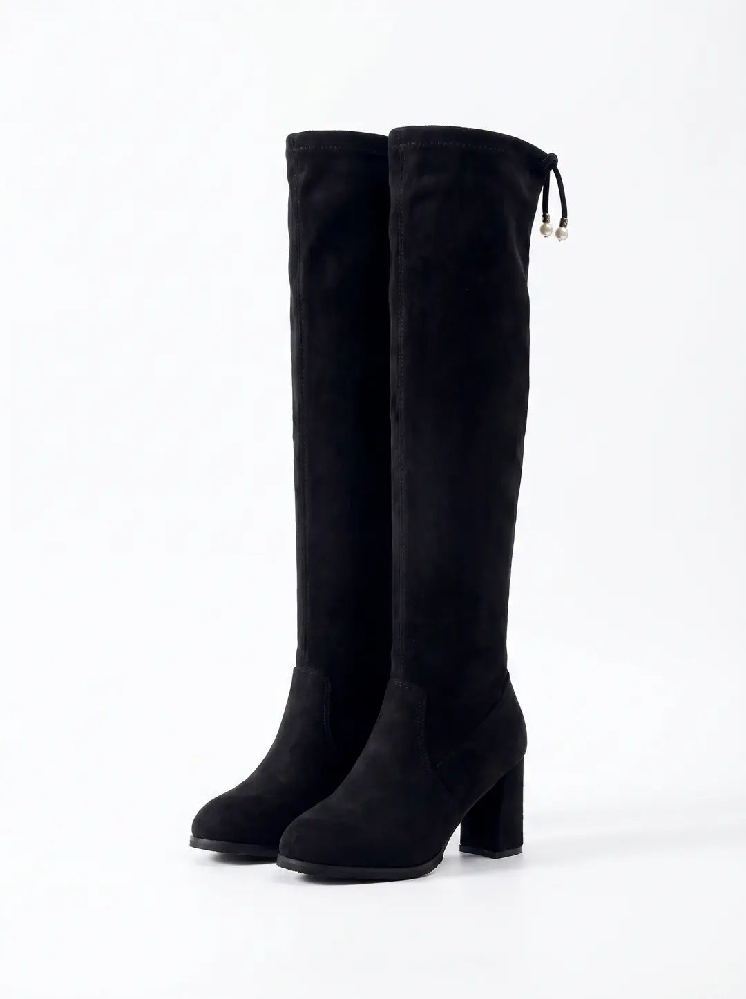 Par de botas negras de ante por encima de la rodilla con tacón grueso. La bota derecha presenta un cordón decorativo con dos perlas blancas en los extremos.