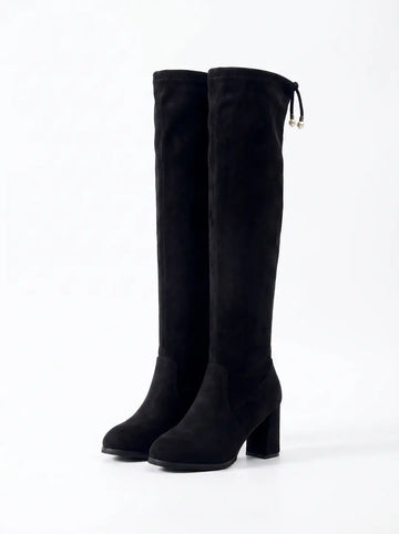 Par de botas negras de ante por encima de la rodilla con tacón grueso. La bota derecha presenta un cordón decorativo con dos perlas blancas en los extremos.