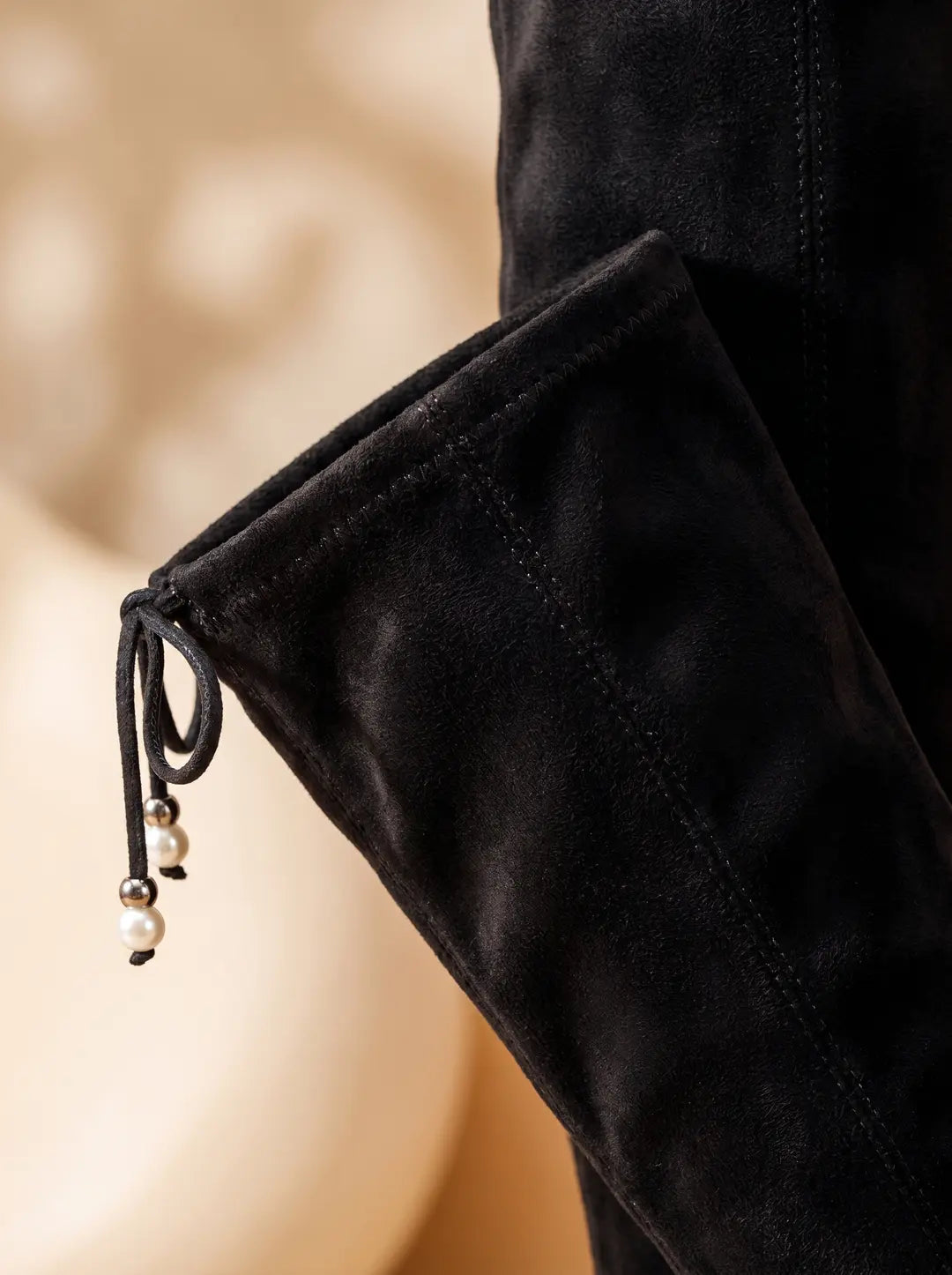 Detalle de la parte superior de una bota alta negra de material similar al ante, con un cordón de ajuste en el borde. El cordón tiene dos cuentas de perlas blancas con detalles metálicos plateados.