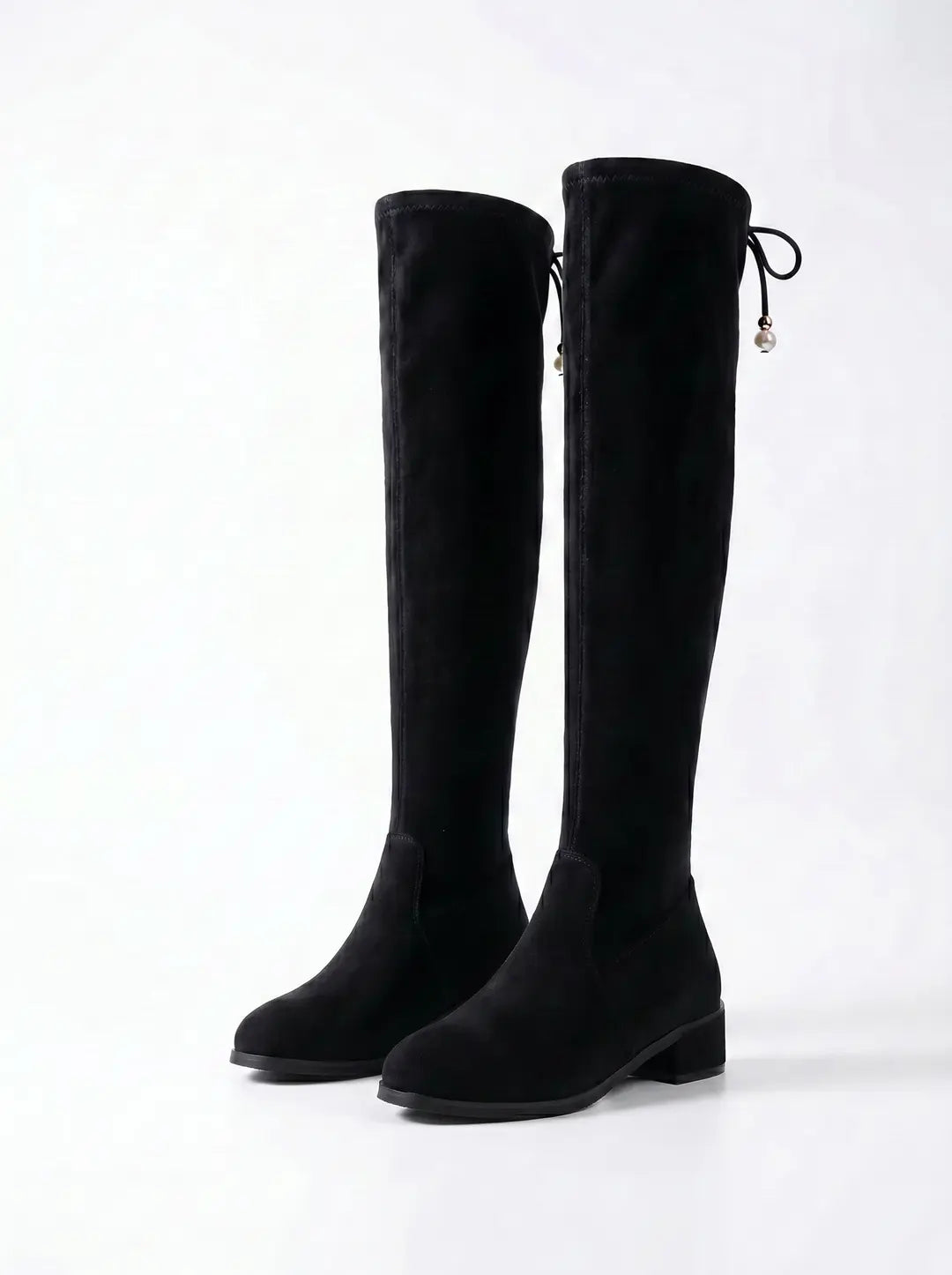 Par de botas negras altas hasta el muslo, con textura similar al ante. Presentan un tacón bajo y un cordón decorativo en la parte superior con abalorios blancos en forma de perla. Fondo blanco.