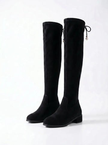 Par de botas negras altas hasta el muslo, con textura similar al ante. Presentan un tacón bajo y un cordón decorativo en la parte superior con abalorios blancos en forma de perla. Fondo blanco.