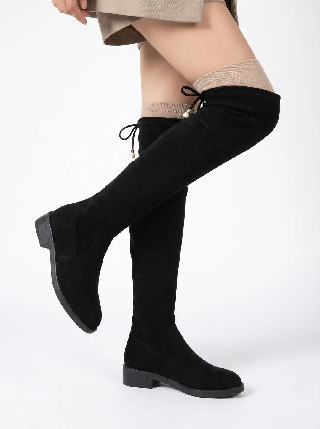Botas negras altas hasta el muslo con apariencia de gamuza, tacón bajo y lazos decorativos con perlas en la parte superior trasera. Se ven sobre medias beige claro.