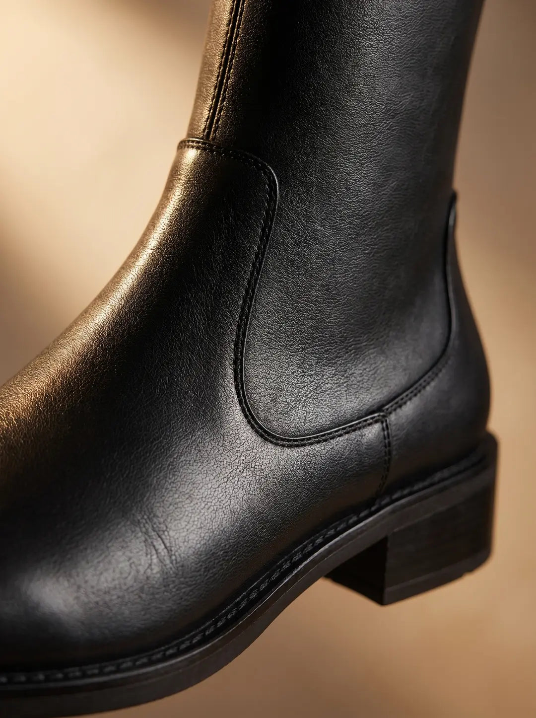 Primer plano de una bota de cuero negra. Se aprecian las costuras detalladas, la textura granulada del cuero y parte de la suela con un tacón bajo.