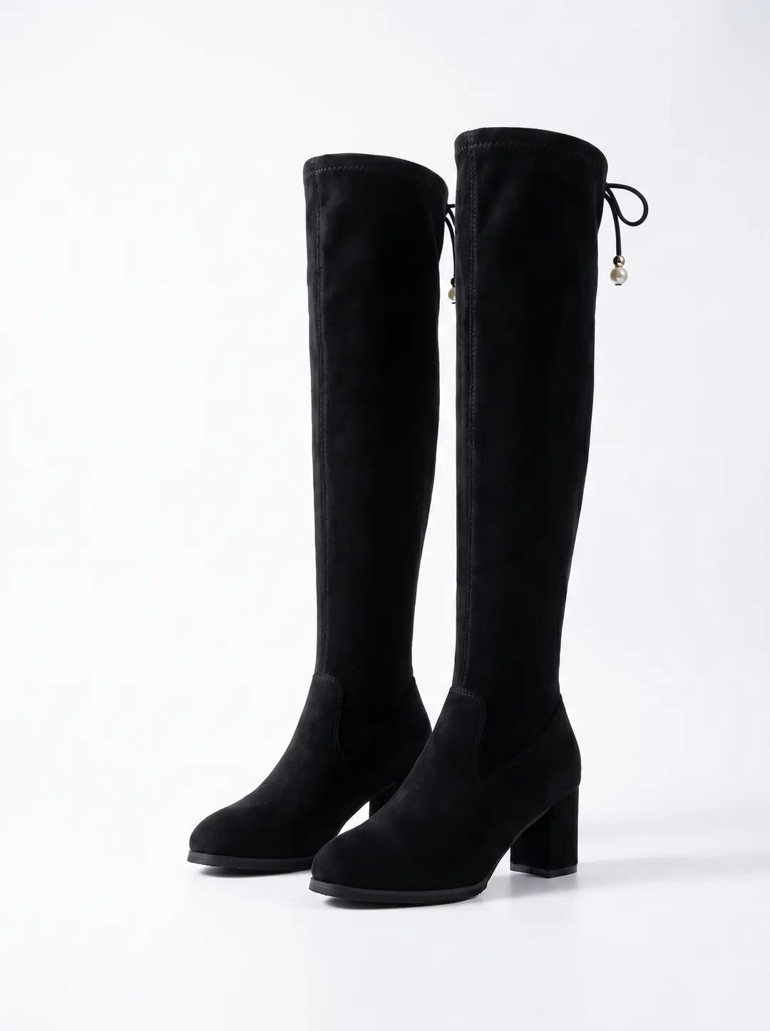 Dos botas altas negras de material similar a la gamuza, con tacones de bloque. Cada bota tiene un lazo decorativo con una perla en la parte superior trasera.