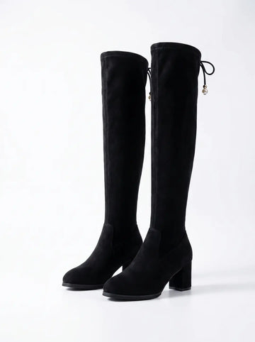 Dos botas altas negras de material similar a la gamuza, con tacones de bloque. Cada bota tiene un lazo decorativo con una perla en la parte superior trasera.