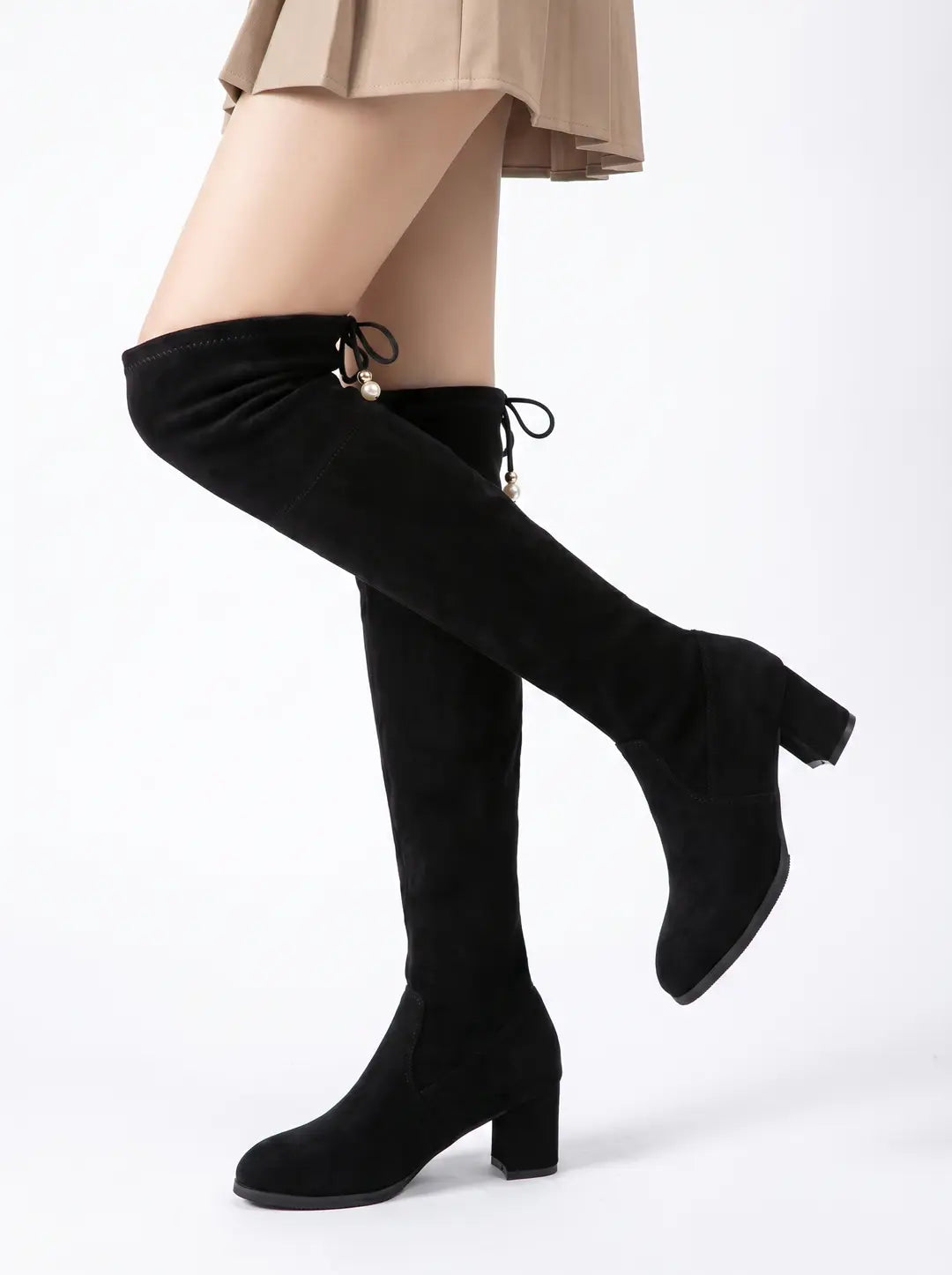 Un par de botas mosqueteras negras de ante con tacón de bloque. La parte superior de las botas presenta cordones decorativos con pequeñas cuentas doradas y perlas. Las botas son llevadas por una persona que también viste una falda plisada de color beige. Fondo blanco.