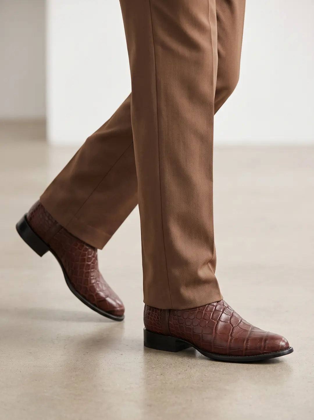 Botas marrones con textura de piel de cocodrilo, suela y tacón negros, combinadas con pantalones marrones, en las piernas de una persona que camina sobre un suelo claro.