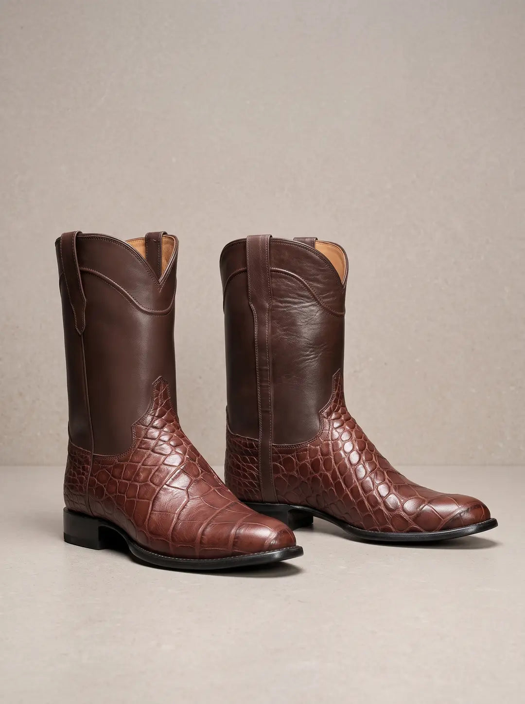 Par de botas vaqueras marrones con la parte del pie en textura de piel de cocodrilo y la caña de cuero liso, sobre una superficie clara.