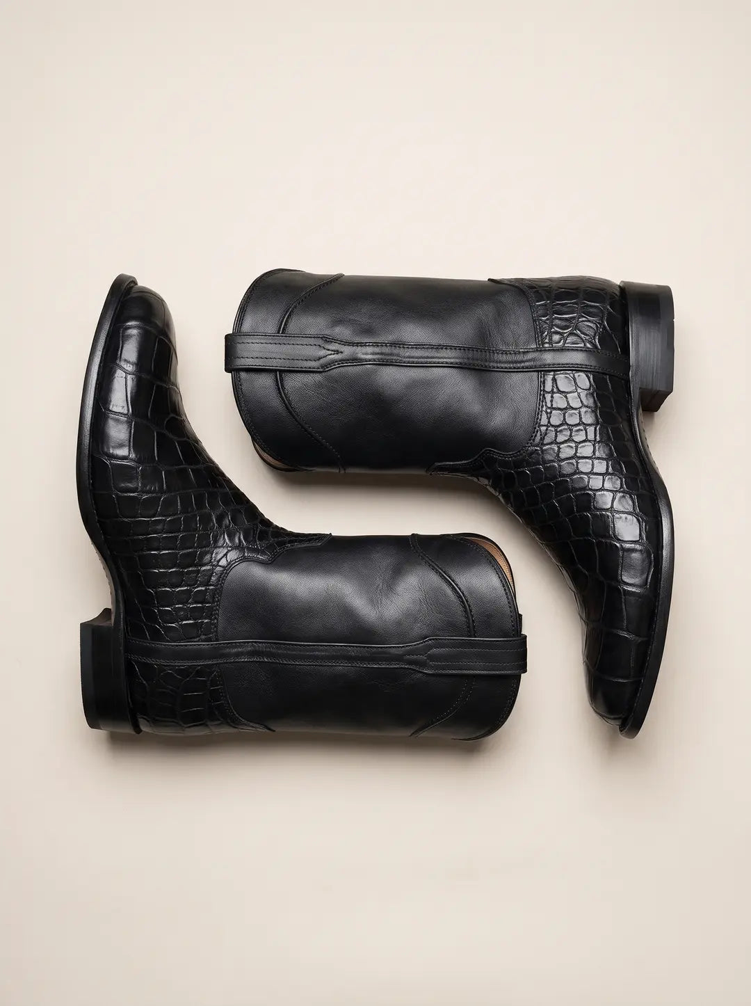 Par de botas de piel negras. La parte inferior, incluyendo la puntera y el talón, presenta un patrón texturizado similar a piel de cocodrilo. La caña es de cuero liso con una tira decorativa. Las botas están colocadas de forma opuesta sobre una superficie beige.