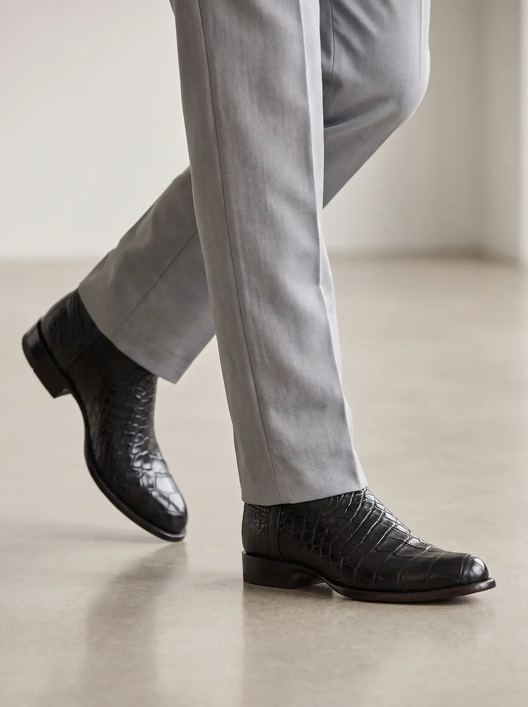 Piernas con pantalones de vestir grises claros y zapatos negros de piel de cocodrilo caminando sobre un suelo de color beige.