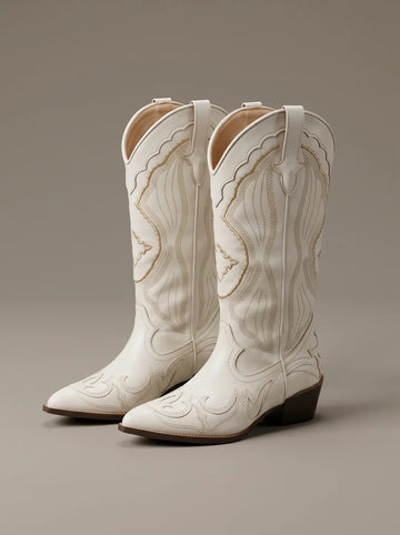 Par de botas vaqueras blancas con puntera puntiaguda y tacón marrón oscuro. Presentan costuras decorativas intrincadas en tono beige que adornan la caña y el empeine.