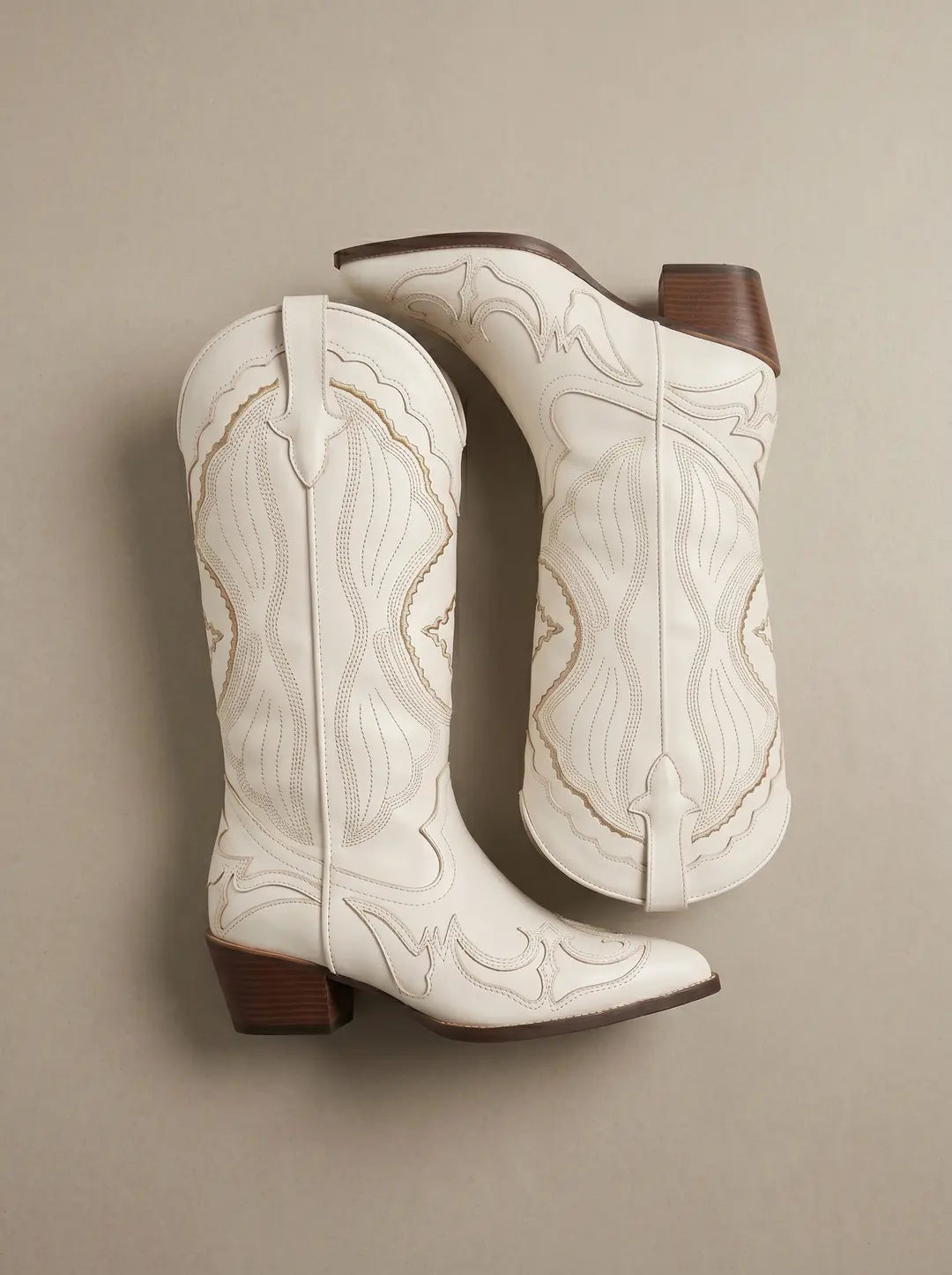 Par de botas vaqueras de color blanco con bordados decorativos. Tienen puntera afilada y tacones apilados de color marrón. Las suelas son oscuras. Están dispuestas sobre una superficie lisa de color beige claro.