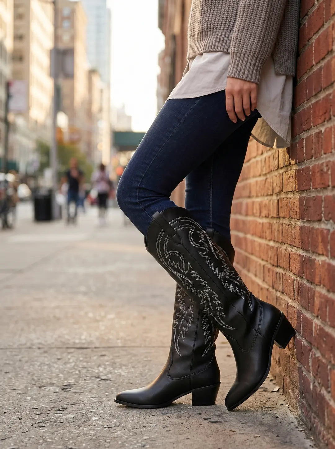 Un par de botas vaqueras negras con bordados blancos, puestas en los pies de una persona que lleva jeans azules, apoyada contra una pared de ladrillo.