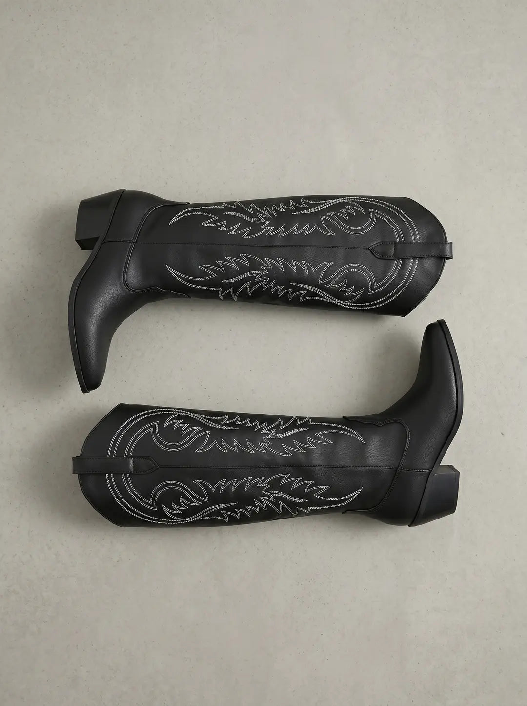Dos botas vaqueras negras con bordados blancos decorativos, dispuestas horizontalmente sobre una superficie gris clara.