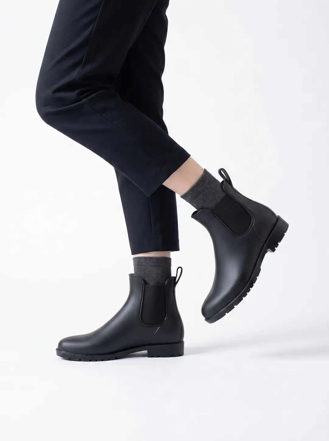 Piernas de una persona con pantalones negros, calcetines grises y botas de lluvia negras estilo Chelsea, una de las cuales está ligeramente levantada. Las botas tienen paneles elásticos laterales y una lengüeta en la parte trasera.
