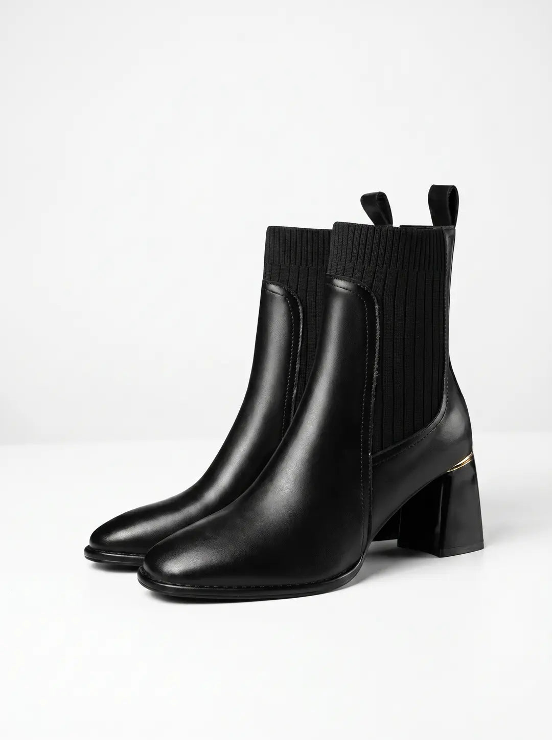 Par de botines negros de mujer con parte inferior de cuero liso y parte superior de tejido elástico acanalado. Tienen tacones de bloque negros con un detalle dorado en la parte superior y presillas traseras.