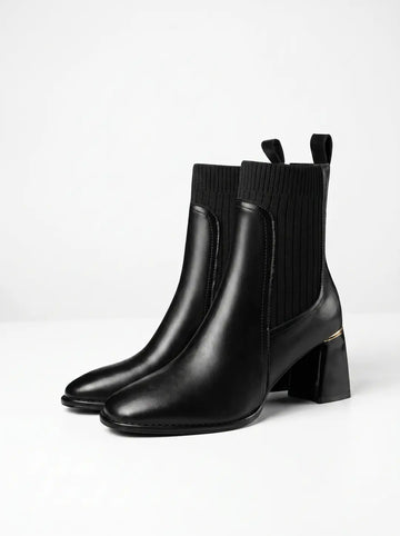 Par de botines negros de mujer con parte inferior de cuero liso y parte superior de tejido elástico acanalado. Tienen tacones de bloque negros con un detalle dorado en la parte superior y presillas traseras.