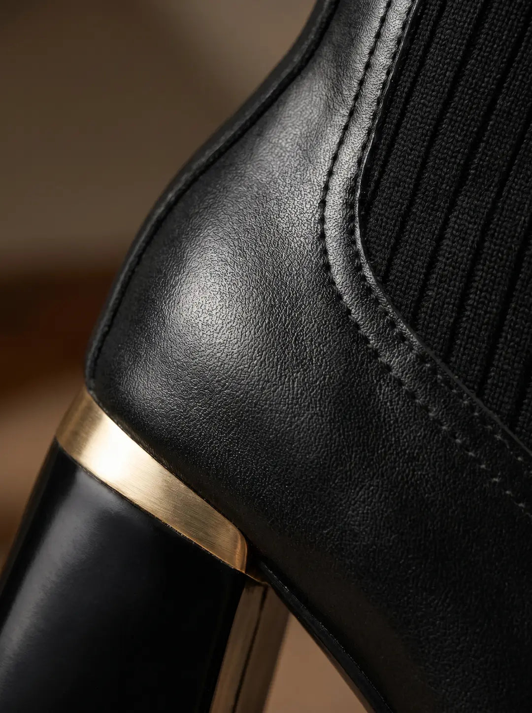 Detalle de una bota negra con textura de piel, un panel lateral de tela acanalada negra y un tacón negro con una banda metálica dorada.