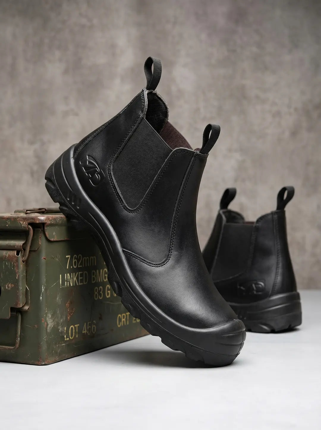 Un par de botas negras estilo Chelsea con paneles laterales elásticos y suelas robustas. La bota delantera está apoyada sobre una caja metálica verde oliva desgastada con inscripciones, y la otra bota se ve parcialmente desenfocada detrás. Ambas botas tienen tiradores en el borde superior.