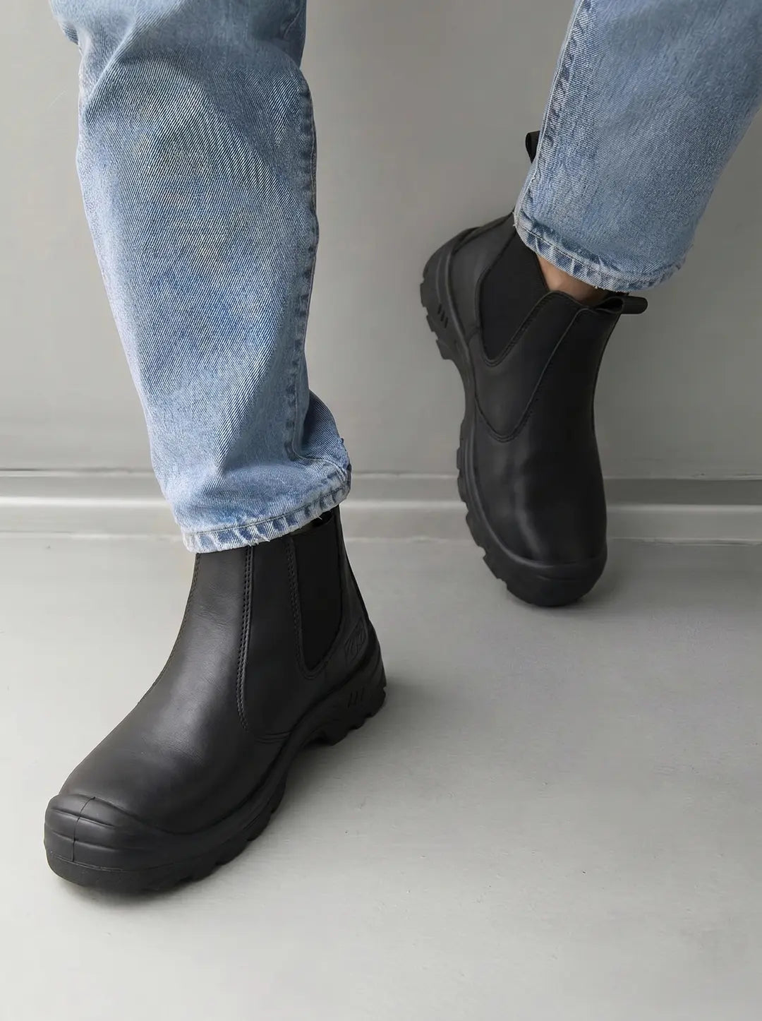 Un par de botas negras estilo Chelsea con suelas gruesas y dentadas, usadas con jeans azules claros, sobre un fondo gris claro.