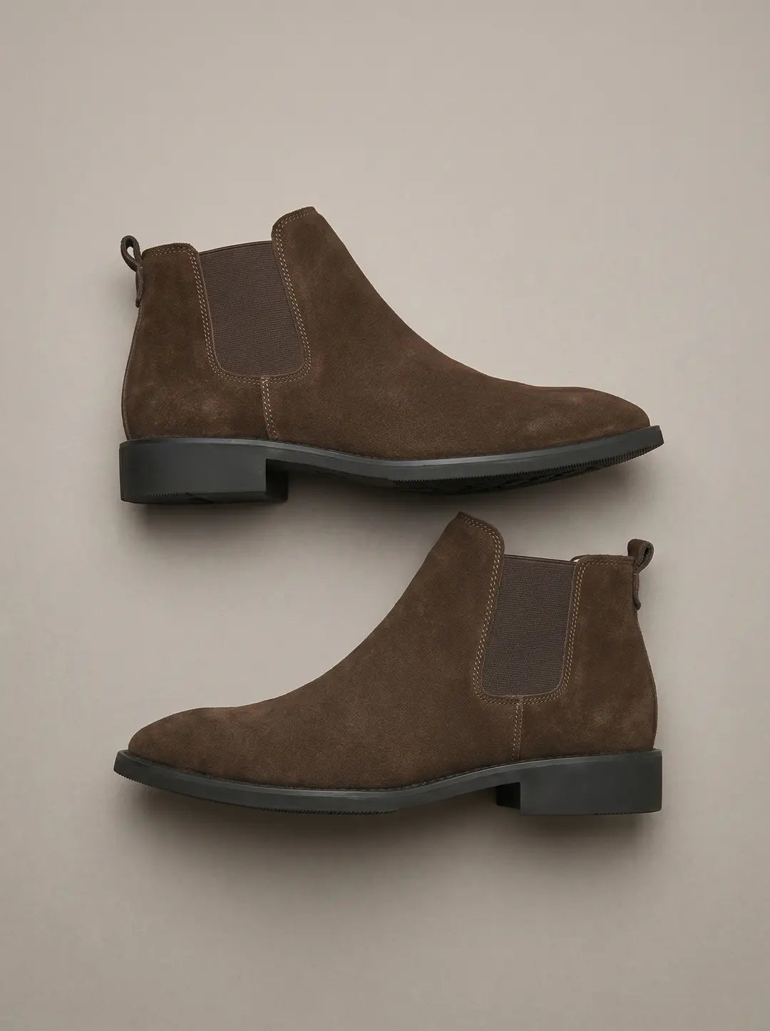 Dos botas Chelsea de gamuza marrón con paneles laterales elásticos y suelas negras, vistas desde arriba sobre un fondo liso.