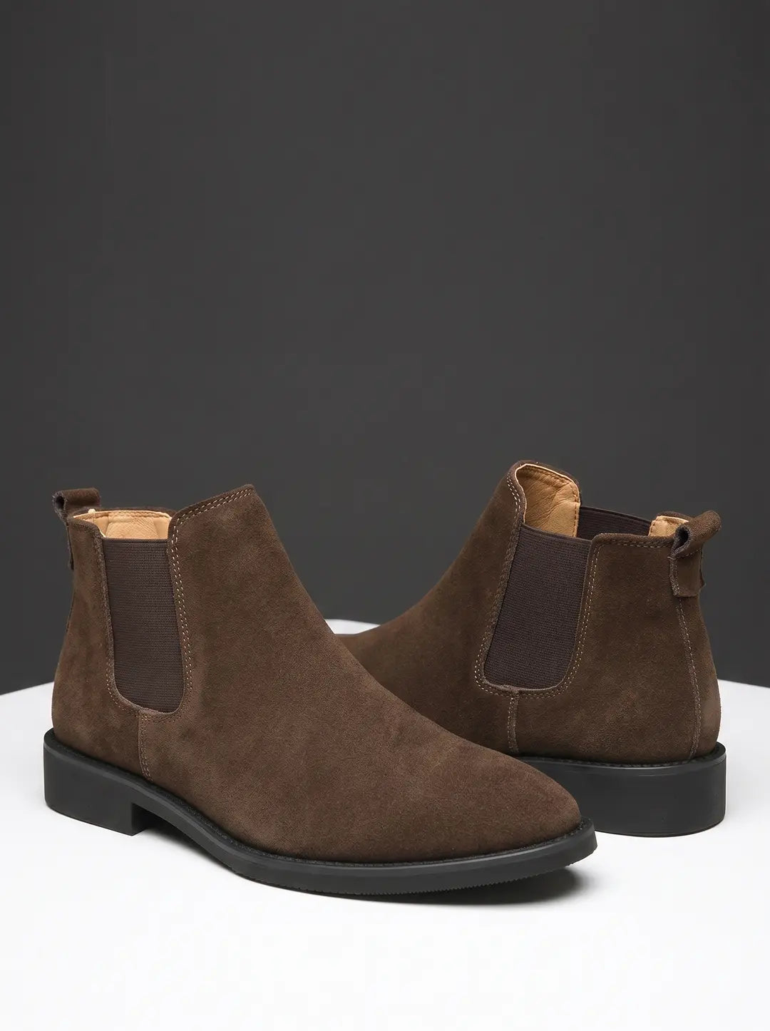 Un par de botas Chelsea de ante marrón con paneles elásticos laterales y suelas negras, exhibidas sobre una superficie blanca contra un fondo gris oscuro.