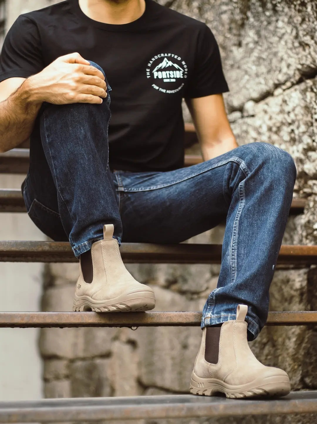 Persona sentada en escaleras metálicas, vistiendo camiseta negra con un logo blanco y jeans azules. Lleva puestas botas tipo Chelsea de color beige claro con suelas robustas, paneles elásticos laterales marrones y una lengüeta trasera.